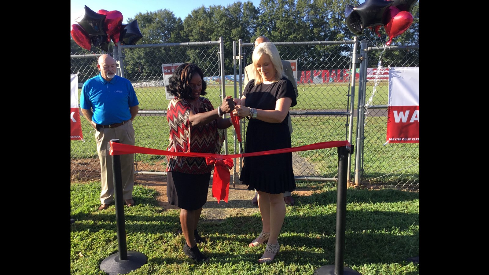 Principal Jones and county representative using red scissors to cut ribbon in front of track fence